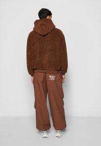 Brown fleece hoodie with a textured finish, paired with loose brown cargo pants featuring logo detail on the back. Grey sneakers complete the look.