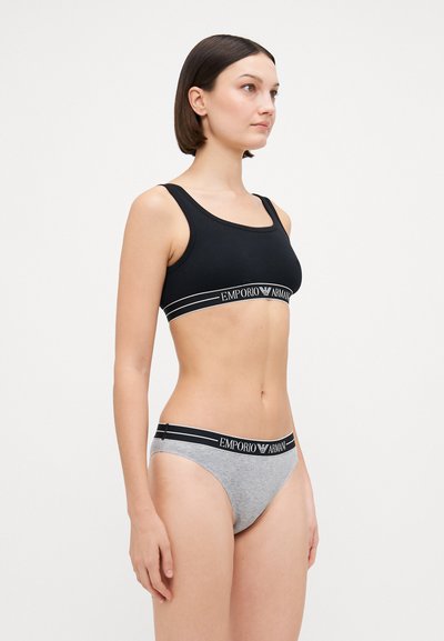Melna bralette ar platām siksnām un pelēkajiem apakšveļas biksītēm ar logotipa jostasvītru, izceļot pieguļošu dizainu un mīksto kokvilnas materiālu.