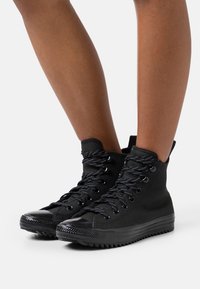 Svart högprofil canvaskängor med en texturerad gummisula, rund tå, kontrasterande snörning och en glänsande tåskydd.