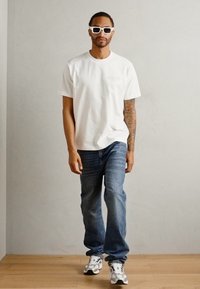 Witte katoenen t-shirt met een klein bedrukt logo, gecombineerd met blauwe spijkerbroeken en grijs-witte sneakers. Het model draagt ook witte zonnebrillen.