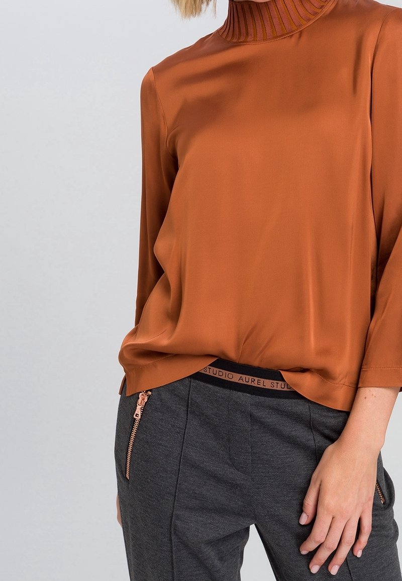 Blouse en soie orange brûlé avec un col montant côtelé, associée à un pantalon gris doté d'une poche zippée et d'une ceinture marquée.