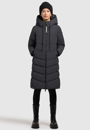 Black padded long coat with a hood, featuring a front zipper, drawstrings, and side pockets. Worn with black leggings and combat boots.