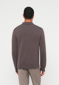 Mann trägt einen braunen Strickpullover mit langen Ärmeln und hellbraune Hose, steht mit dem Gesicht zu einer schlichten weißen Wand und die Arme locker an den Seiten.