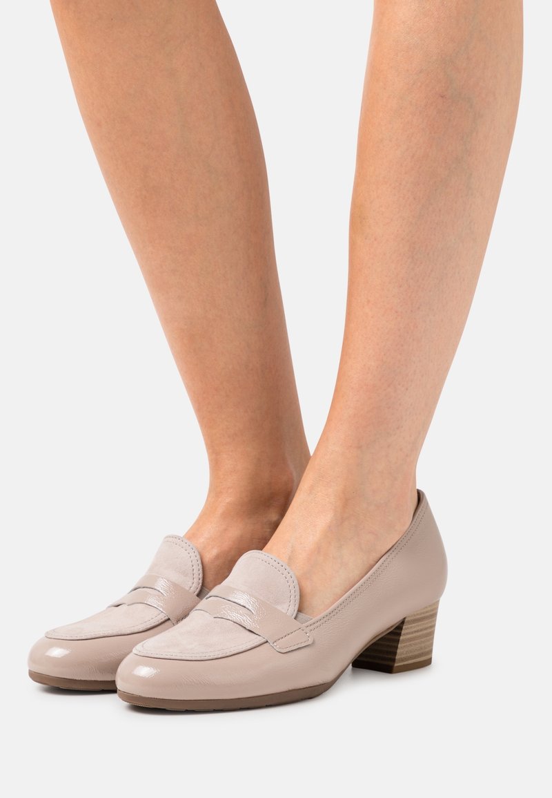 Mocassins à talons bas en cuir beige et daim, portés pieds nus, avec un talon en bois empilé, sur un fond uni.