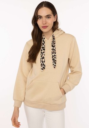 Frau trägt beigen Hoodie mit Leopardenmuster-Kordeln und weiße Hose, steht mit einer Hand in der Tasche vor weißem Hintergrund.