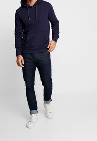 Granatowa bluza z kapturem z przednią kieszenią i akcentami w formie sznurka, zestawiona z ciemnymi dżinsami i białymi trampkami za kostkę.