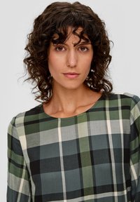 Langärmliges Karo-Kleid mit grünen, schwarzen und beigen Karomustern. Weicher Stoff mit Rundhalsausschnitt und leicht gepufften Ärmeln.
