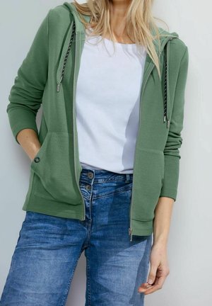 Femme portant un sweat à capuche vert zippé, un t-shirt blanc, et un jean bleu taille haute avec fermeture double bouton.