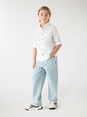Giovane in piedi con una mano in tasca, indossa una camicia bianca con bottoni, jeans azzurri e sneaker bianche e nere su sfondo semplice.