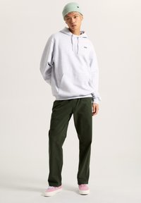 Felpa grigia con tasca frontale, berretto verde, pantaloni verde oliva e sneakers rosa, in piedi contro uno sfondo neutro.