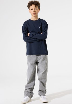 Camisa de manga larga navy con texto blanco, combinada con pantalones vaqueros sueltos gris claro y zapatillas blancas. El conjunto tiene un ajuste relajado.