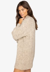 Beżowy, oversize sweter o miękkim, teksturowanym wykończeniu, długości sięgającej do połowy ud, z długimi rękawami, wysokim kołnierzem i minimalnymi szwami.