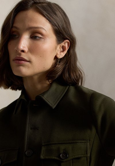 Olijfgroene overhemdjas met een kraag, twee voorzakken en zwarte knopen. Gouden oorringen en een gladde textuur.