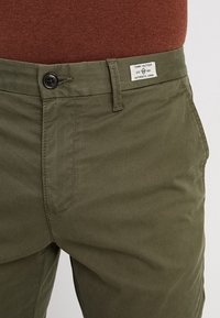Pantaloni chino verde oliva realizzati in tessuto leggero, con chiusura anteriore a bottoni, linee pulite e etichetta sulla vita.