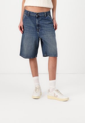 Person iført knælange blå cowboyshorts, hvide sneakers med beige accenter, hvide crew-sokker og en cropped hvid sweater.