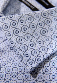 Camicia con colletto in tessuto azzurro chiaro con un motivo floreale ripetuto in blu scuro, che presenta una texture morbida e un design strutturato.