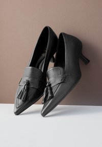 Chaussures à talons hauts noires en cuir, avec un bout pointu, des pampilles décoratives et un petit talon empilé. Texture lisse avec des détails de couture.