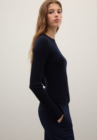 Pull côtelé marine à manches longues et col rond, associé à un pantalon foncé. Le tissu semble lisse avec une coupe ajustée.