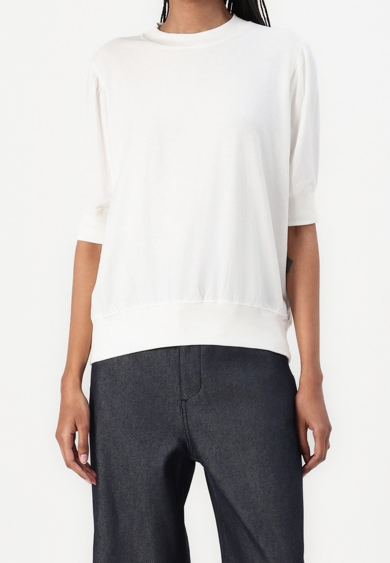 Lätt, krämfärgad skjorta med rund halsringning, trekvartslånga ärmar och rynkad nederkant, matchad med mörka denimbyxor.