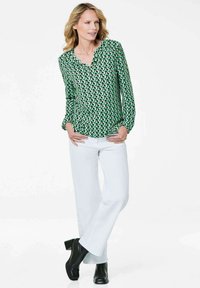 Blusa verde con motivo e dettagli neri, scollatura a V, maniche lunghe. Abbinata a pantaloni larghi bianchi e stivali neri con tacco.