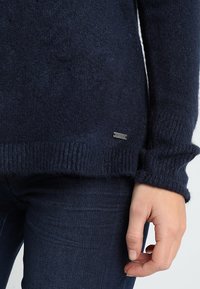 Marinblå ulltröja med ribbad nederkant och muddar. Har en liten metall-logotagg nära nederkanten. Bärs över mörka denimjeans.