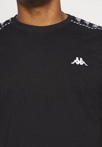Černé tričko s krátkým rukávem a kulatým výstřihem. Obsahuje bílé logo Kappa a vzorované detaily na ramenou. Vyrobeno z hladké tkaniny.