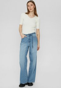 Weißes, geripptes Kurzarmoberteil mit gewelltem Halsausschnitt, kombiniert mit hellblauen, weit geschnittenen Jeans, die eine Kordel und Fronttaschen besitzen.