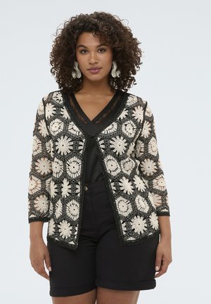 Vrouw met krullend haar die een witte gebreide cardigan met bloemenmotief draagt over een zwarte top en zwarte short, staand tegen een lichte achtergrond.
