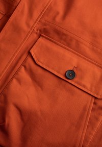 Chaqueta de tejido naranja con un bolsillo en el pecho con botón; textura suave; presenta un pespunteado y un detalle de botón negro en el bolsillo.