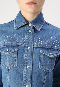 Camicia di denim con lavaggio blu, caratterizzata da borchie argentate sul petto, due tasche anteriori, colletto a punta e chiusura con bottoni a pressione.
