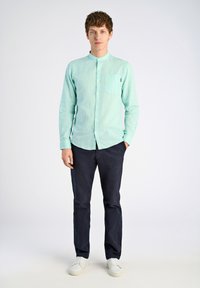 Vaalean minttuvihreä pitkähihaisella paidalla, jossa on nappikaulus ja rintatasku, yhdistettynä tummansinisiin housuihin ja valkoisiin lenkkareihin. Sujuva pinta.