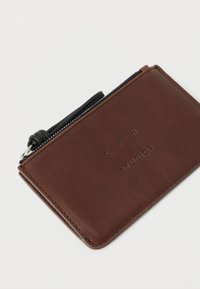 Brązowy skórzany portfel na karty z czarnym uchwytem na suwak oraz wytłoczonym logo "CPH STUDIOS" z przodu, pochylony na białym tle.