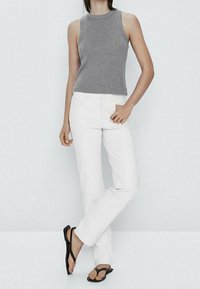 Woman standing with one hand in white pants pocket, wearing a sleeveless gray top and black thong sandals against a plain background.