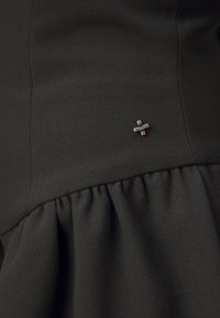 Črna obleka iz tkanine z nabranim volanom na dnu in srebrnim kovinskim dodatkom na strani, ki poudarja njen strukturiran dizajn.