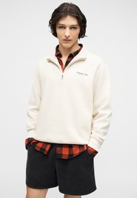 Νέος άντρας με λευκό φούτερ με φερμουάρ στο στήθος πάνω από κόκκινο και μαύρο πουκάμισο καρό, με μαύρες βερμούδες και τα χέρια στις τσέπες, στέκεται μπροστά σε λευκό φόντο.