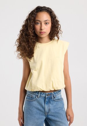 Jeune femme aux cheveux bouclés portant un haut sans manches jaune pâle et un jean taille haute bleu, debout devant un fond blanc uni.