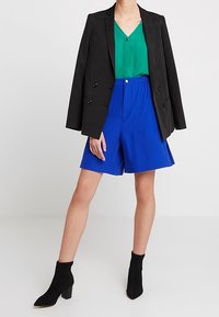 Svart dubbelknäppt kavaj, grön blus med knapp, blå skräddarsydda shorts och svarta ankelskor med blockklack.