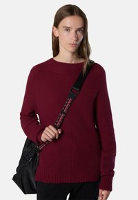 Maglione a maglia bordeaux con collo a girocollo a coste e maniche lunghe, caratterizzato da un motivo testurizzato. Una borsa nera con manici intrecciati è tenuta in mano.
