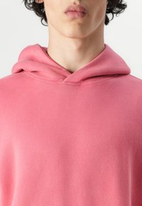 Hoodie rose en tissu doux, avec une poche kangourou à l'avant et une capuche avec cordon de serrage, présentant des détails de couture visibles.