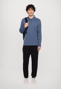 Langærmet blå polo med lynlås krave, parret med sorte cargo bukser. Modellen holder en sort rygsæk og har hvide sneakers på.
