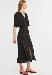 Zwarte midi-jurk met fluttermouwen, voorzien van een kersenpatroon. Diepe V-hals en een zijsplit. Gepaard met rode sandalen met bandjes.
