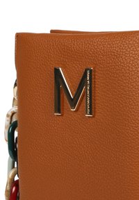 Hnědá kožená taška přes rameno s výrazným kovovým logem "M" a barevnými řetězovými akcenty na jedné straně. Viditelný texturovaný povrch.