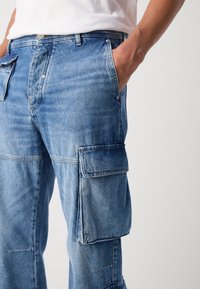 Pantalones cargo de color azul claro hechos de mezclilla. Cuentan con dos bolsillos laterales y un corte relajado. Costuras visibles en las juntas y una cintura con botón.