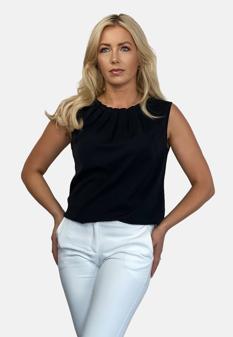 Czarna bezrękawnikowa bluzka z plisowanym dekoltem, w połączeniu z białymi dopasowanymi spodniami, gładka tekstura i dopasowany krój. Neutralne tło.