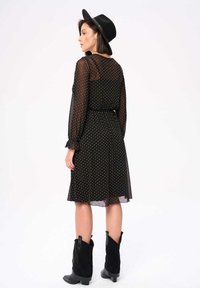 Zwarte, doorschijnende jurk met stippen, lange mouwen en gerafelde manchetten, een aansnoerende taille, een vloeiende rok, gecombineerd met zwarte cowboylaarzen en een brede hoed.