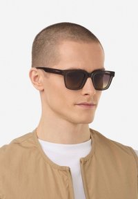 Occhiali da sole neri con montatura quadrata, lenti sfumate marroni, finitura testurizzata e un logo minimale sulla tematica. Indossati da un modello.