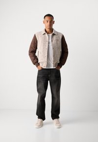 Giovane uomo in giacca imbottita beige con maniche marroni, t-shirt bianca, jeans neri e sneakers beige chiaro, in piedi con le mani in tasca.