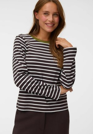 Femme aux longs cheveux bruns souriante, portant une chemise à manches longues rayée noir et blanc avec un col vert olive, posant avec une main près de son cou.