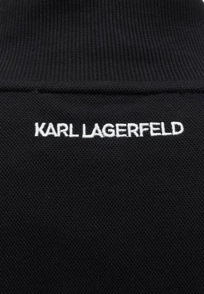 Tissu noir texturé avec inscription brodée blanche "KARL LAGERFELD" sous une section de col côtelé.