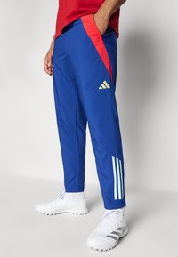 Pantalones atléticos azules con acentos rojos a los lados, que presentan tres rayas blancas en la pierna izquierda. Hechos de un material suave y ligero.